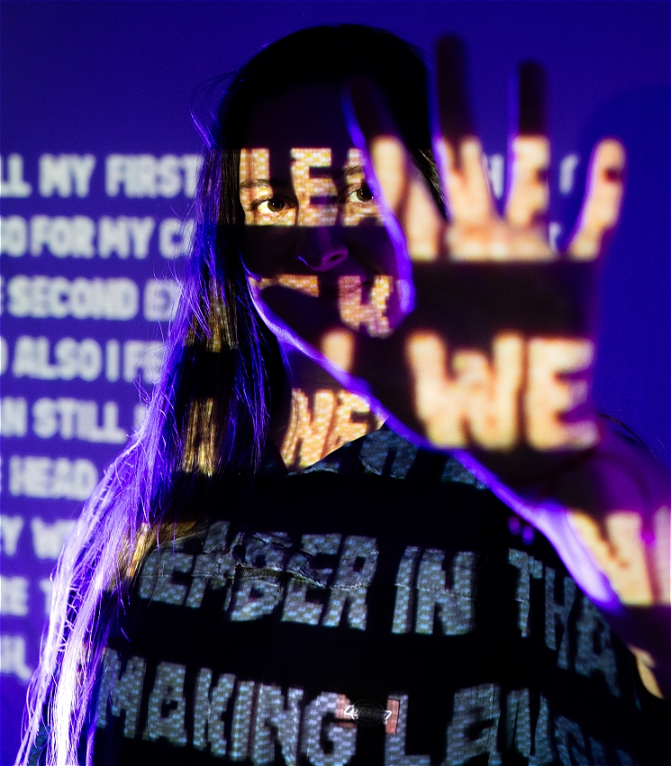 The image shows a woman with text projected onto her face and body. She has her hand raised, partially obscuring her face, with words visible on her hand and shirt. The background is dark, with more text projected, creating a visually striking and mysterious scene.