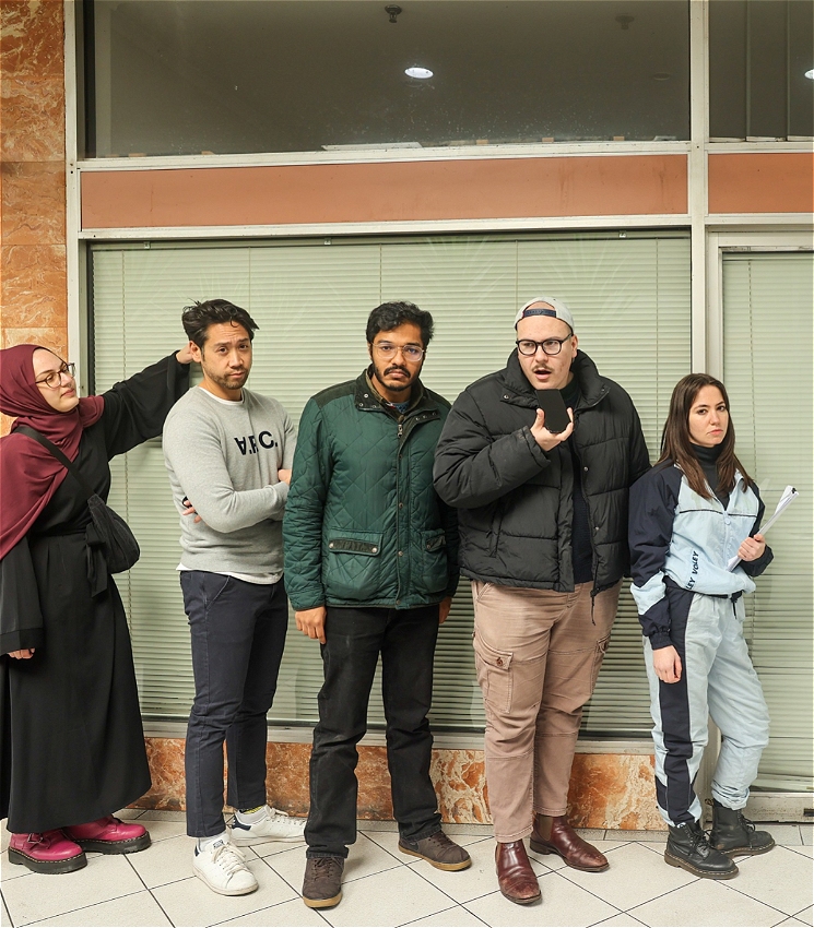 Five people (two women and three men). The group is standing in front of a glass door and windows with blinds, part of a building with a marble exterior. The overall atmosphere suggests they are waiting or posing outside an official government office.