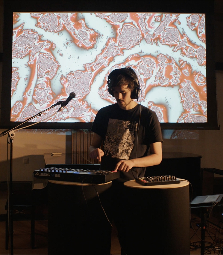 A man is standing in front of a microphone, immersed in playing electronic music equipment with his one hand pressing a key, while the other is captured hovering over. He is wearing headphones and a dark shirt. Behind the man is a visual projection of pink and grey abstract patterns.