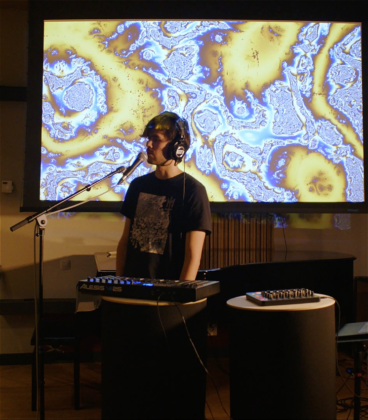 Side profile of a man speaking into a microphone, standing behind electronic music equipment. He is wearing headphones and a dark shirt with white print. Behind the man is a visual projection of blue and yellow abstract patterns.