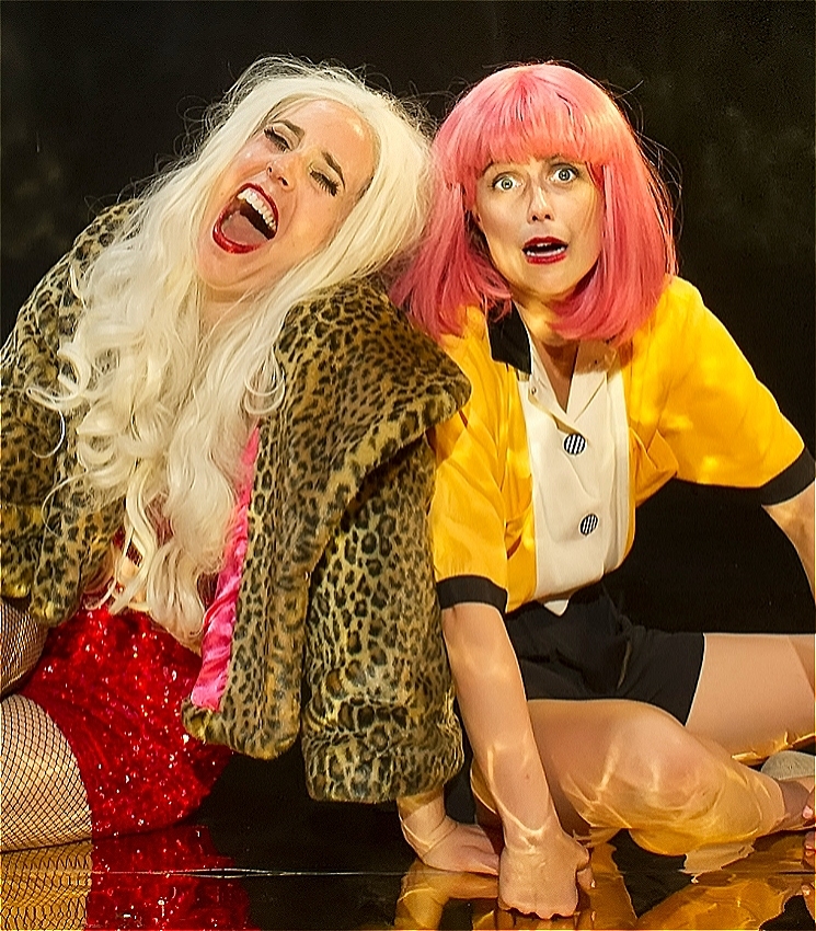 The image is a portrait of two women sitting on the floor against a black background. The women on the left is caucasian, wearing a long blonde wig, a leopard print coat, and is laughing with her eyes closed and head tilted back. The woman on the right is caucasian, wearing a yellow short-sleeved shirt, a pink bob wig, and is making a comically terrified expression towards the camera.