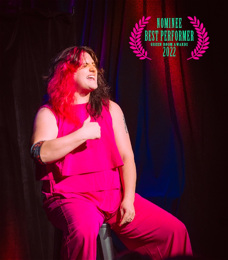 An image of Tricksy seated on black stool on stage, under a white spotlight. Tricksy is wearing a bright pink outfit, has Red and black hair, and her eyes are closed, as she passionately points at her chest with her right hand. Her left arm rests by her side. In the top right corner, is an award laurels that reads 