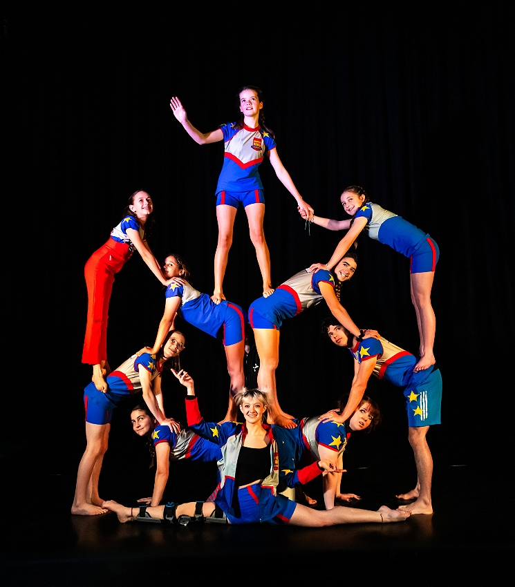 The image showcases a group of young acrobats performing a human pyramid. They are dressed in vibrant blue and red uniforms with star motifs, giving the scene an energetic and patriotic feel. The formation is well-structured, with performers carefully balanced on each other's shoulders and backs. The background is dark, which makes the brightly lit performers stand out even more.