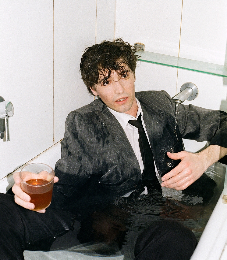 The image depicts a person sitting in a bathtub while fully dressed in a wet suit and tie. They have disheveled hair and are holding a drink in one hand, looking somewhat dazed or contemplative. The setting is a plain, tiled bathroom, and water is still running from the faucet, adding to the chaotic, perhaps melancholic atmosphere. The overall vibe suggests a dramatic or surreal moment, with a touch of dark humor or cinematic intensity, reminiscent of a scene from a French film.