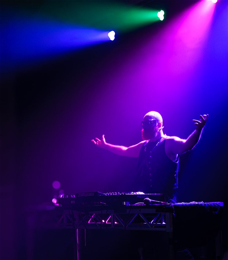 A bald man with a bright red beard stands on stage behind a DJ setup, arms outstretched in a commanding pose. He wears a black vest and a black mask that covers his eyes. The stage is bathed in vibrant purple, pink, and blue lights, creating a dramatic and intense atmosphere.