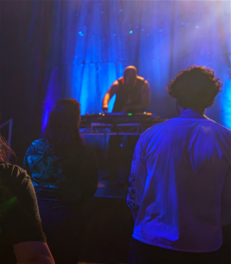 A DJ with a bald head and muscular build stands behind a DJ setup on a dimly lit stage, illuminated by deep blue lights. The DJ's face is slightly obscured as they focus on their equipment. In the foreground, audience members are seen from behind, watching the performance. The scene has a moody, immersive feel, with the blue lighting creating a sense of depth and drawing attention to the DJ as the focal point.