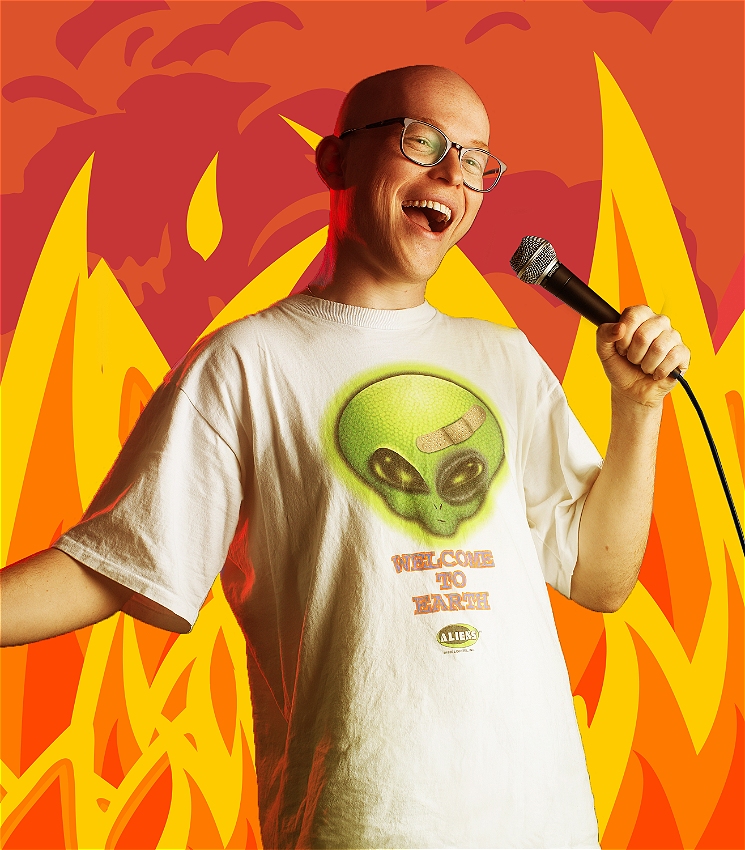 A young man is onstage holding a microphone. He is dressed in a baggy tshirt with an alien on it and looks out with a mischievous smile as he tells a joke.