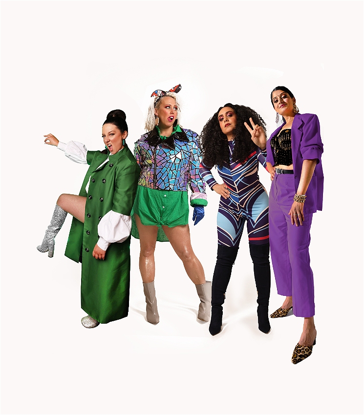 Four women are standing in front of a white background, their legs and feet are on display. They are all wearing different outfits. The woman on the left is wearing a green coat and white boots. The next woman is wearing a colorful jacket, a green skirt, and gray boots. The next woman is wearing a black and white jumpsuit and black boots. The last woman on the right is wearing a purple suit and leopard-print shoes.