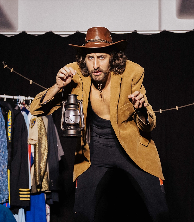 The image shows a man in a theatrical setting, performing in front of an audience. He is wearing a brown cowboy-style hat and a tan blazer with black clothing underneath. He holds a lantern in one hand and appears to be in the middle of an expressive gesture, leaning forward towards the audience. Behind him, there is a clothes rack with various colorful costumes, suggesting a backstage or performance environment. The background is a black curtain, adding a dramatic, focused atmosphere to the scene.