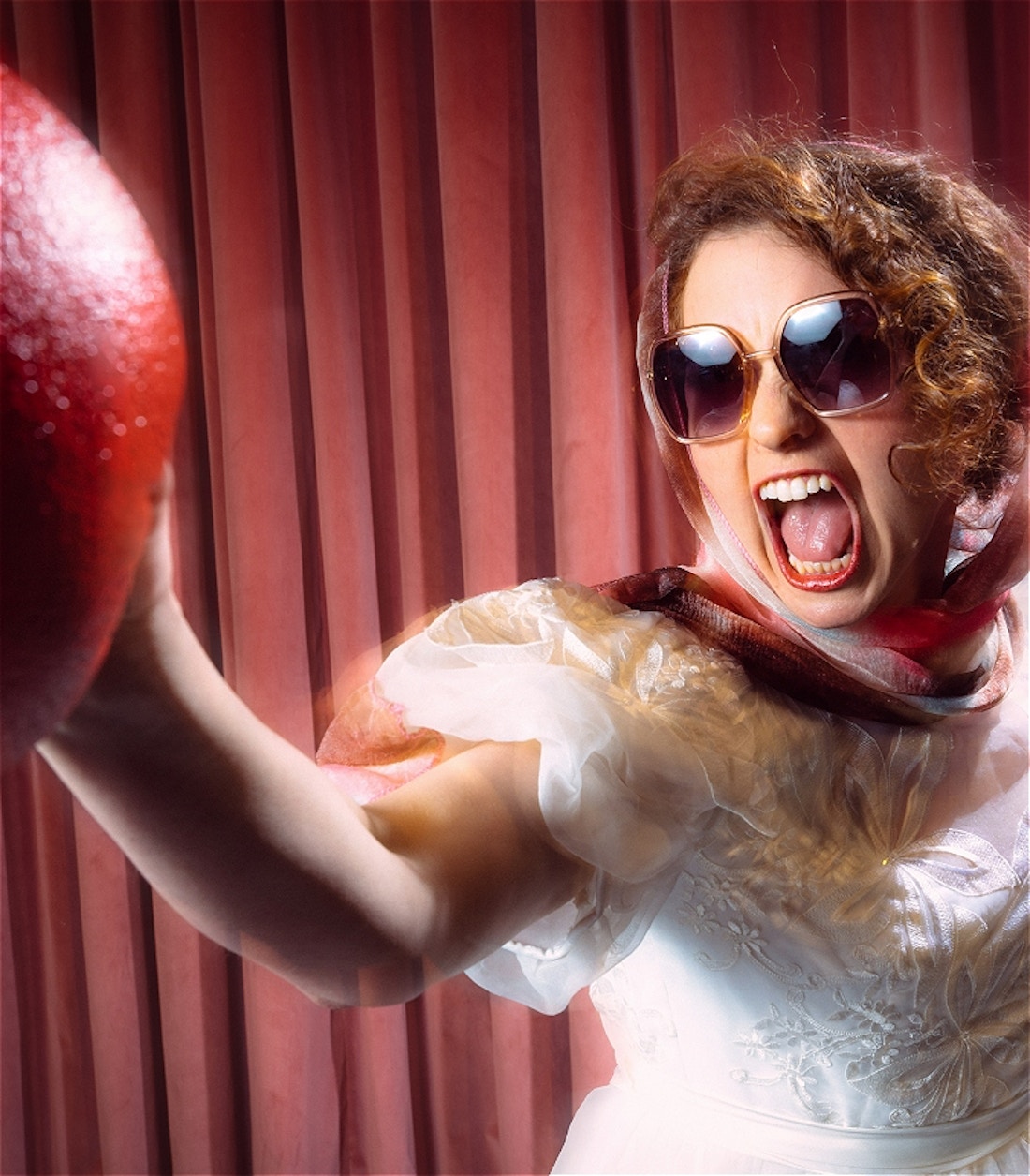 A white woman wearing a white wedding dress, a pastel pink and red head scarf, and big dark sunglasses, has her mouth wide open as if bellowing a war cry. She has her right arm extended in front of her, holding a red dodgeball, as if she is in the process of throwing it.