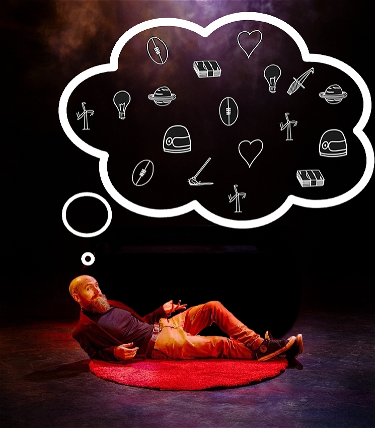 A man with a bald head and full beard is lying on his side on a red circular rug in the center of a dark stage. He appears relaxed, with his legs stretched out and his upper body slightly propped up by one elbow. Above his head is a large white thought bubble filled with whimsical line drawings, including symbols like light bulbs, hearts, helmets, planets, and tools. These images suggest a variety of ideas, thoughts, or inventions.