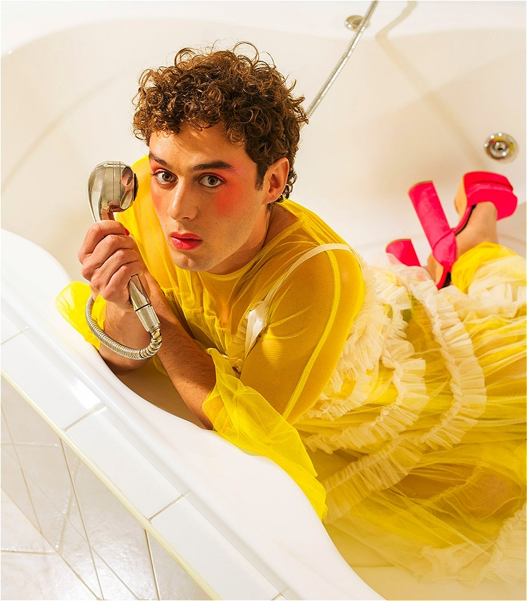 The image shows performer Tomáš Kantor with a curly brown hair, bright pink blush, and lipstick, lying in a white bathtub. They are wearing a sheer yellow outfit with frilled layers and bold pink high heels. The person is holding a showerhead close to their face while resting on their side.