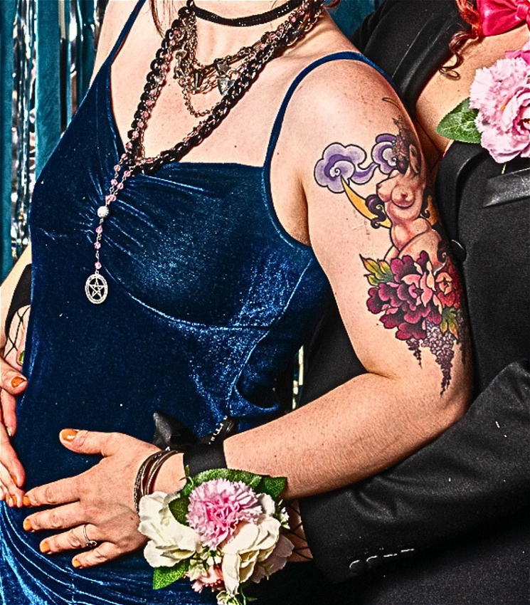 This cropped image shows a close-up of two individuals standing together, focusing on their torsos and arms. The person on the left wears a dark blue velvet spaghetti-strap dress, accessorized with layered necklaces, including one with a pentagram pendant. The other person, positioned slightly behind, is wearing a black jacket with a pink floral corsage pinned to their lapel.