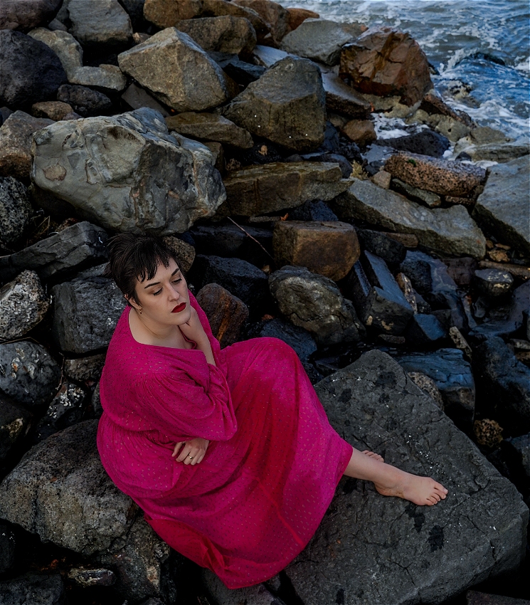 An image from above of Ilana Charnelle, a white woman with short brown hair and red lipstick, sitting on a pile of rocks that leads to the ocean. She is wearing a long pink dress that is wet in patches, and is looking down with a sad expression on her face.