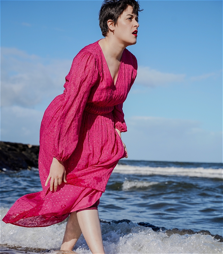 An image of Ilana Charnelle, a white woman with short brown hair and red lipstick, walking in the shallow waves of a beach. The ocean can be seen behind her. She is wearing a long pink dress that she is holding up with her hands to stop the hem falling in the water, and she is looking away from the camera with a bewildered expression.