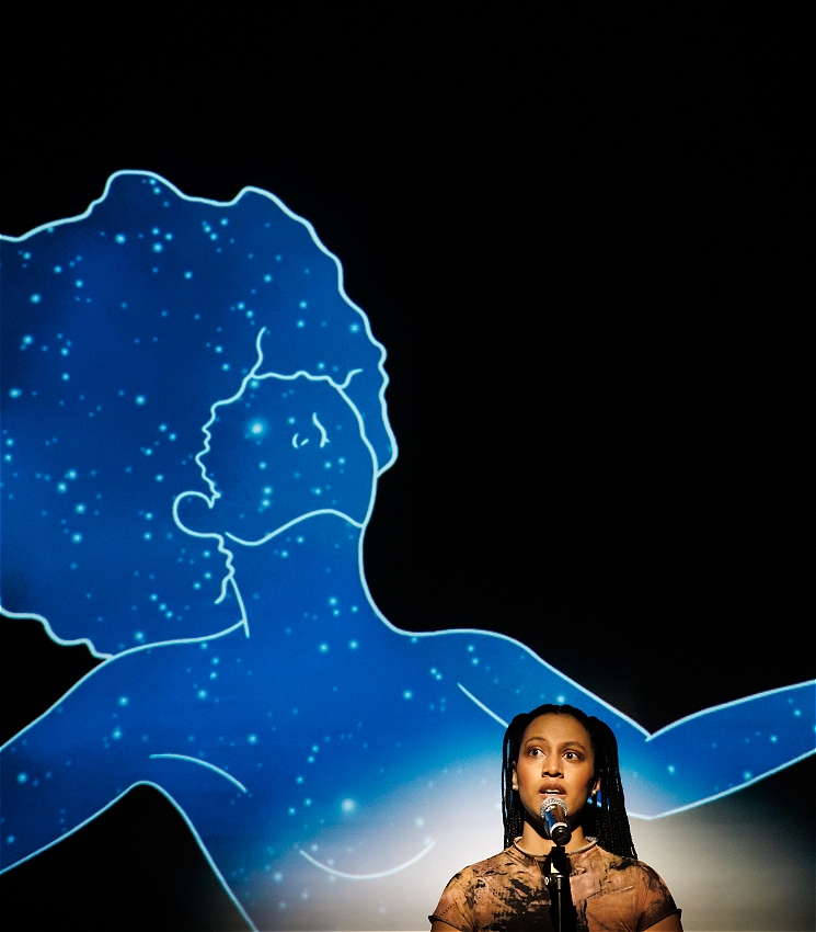 A performer standing on stage with a microphone and stand. Behind her is a projection of a silhouette of a black lady floating in space, she has the texture of a stars
