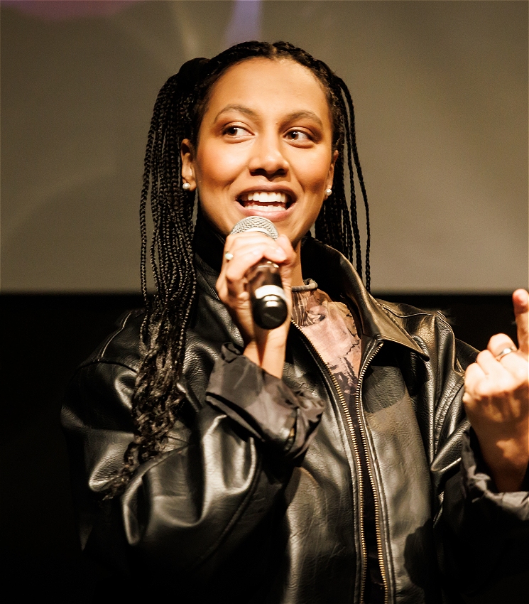 A performer with braids and a black leather jacket smiles as she is talking into a hand held mic.
