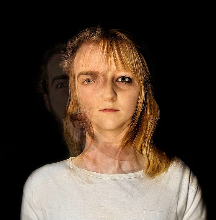 A woman in a white shirt stares bleakly at the camera. A mans face is overlayed over the right side of her face, they appear to be blending together. Another version of the mans face leans out from right side of the head, he is screaming.
The background of the image is black.