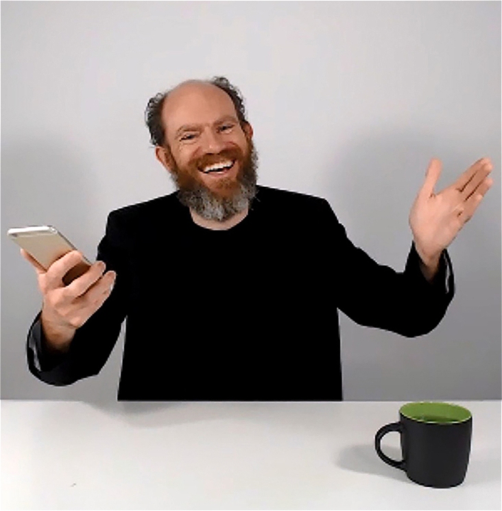A white male with a full reddish-brown beard with gray at the bottom and a bald spot atop his head with dark brown hair on both sides is seated in a chair behind a white desk with both arms raised and a big smile with teeth. He is dressed in an all-black suit. In his left hand is a mobile phone. On the table in front of him is a black mug with a green interior.