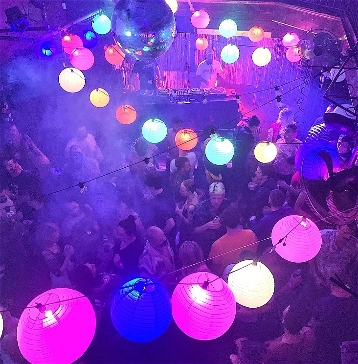 A busy dancefloor with a mirror ball and colourful lanterns above. A DJ plays in the distance.