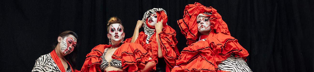 Five performers are posing in zebra print costumes featuring large red frills. Their face paint is white with animalistic details.