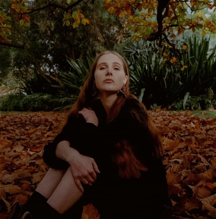 Bec Sykes sits amongst some orange autumn leaves under a tree. She has long red wavy hair and wears a black dress, Blundstone boots and large butterfly earrings. She looks directly into the camera and her gaze is relaxed yet powerful.
