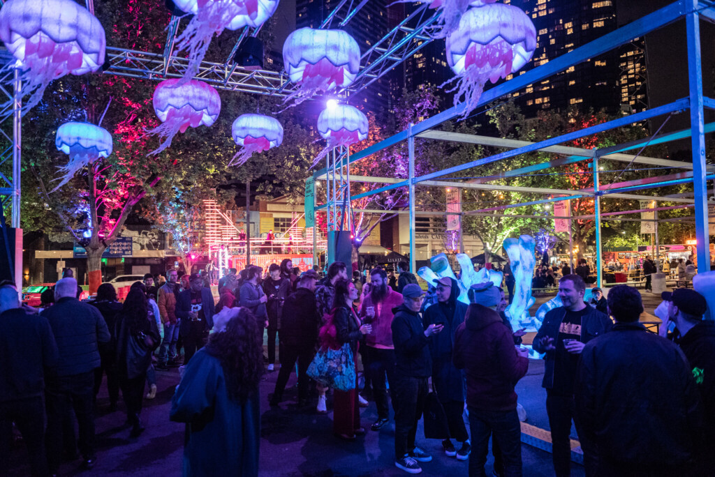 People queue outdoors waiting to go into a show under purple lighting fixtures that look like jellyfish