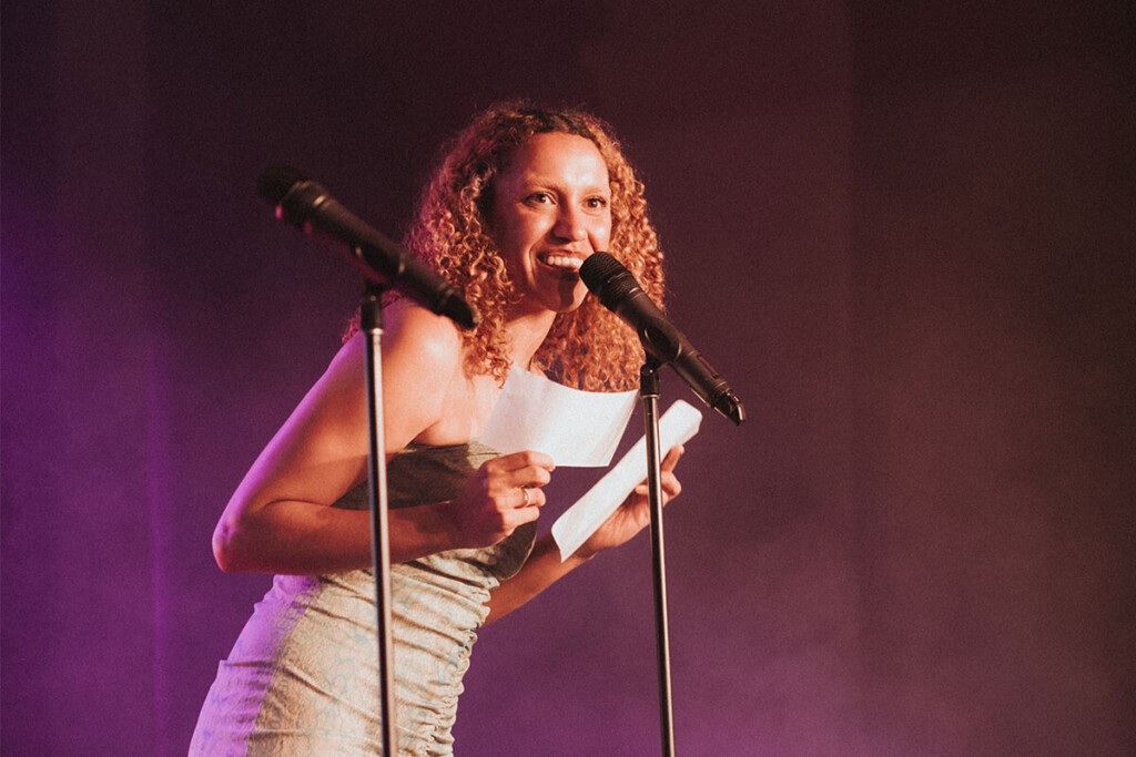 A woman with curly hair stands on stage smiling and reading from a piece of paper into a microphone