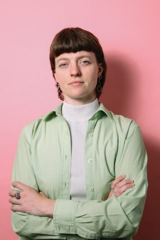 A portrait of Milly Cooper, Access Fringe Program Manager, standing against a solid pink background. Milly has short brown hair with bangs, wears silver hoop earrings, a white turtleneck, and a light green button-up shirt. Their left hand rests on their hip, and they wear a silver ring on one finger. A purple curved banner at the bottom contains bold white text that reads 'MILLY COOPER' and smaller white text below that reads 'ACCESS FRINGE PROGRAM MANAGER'